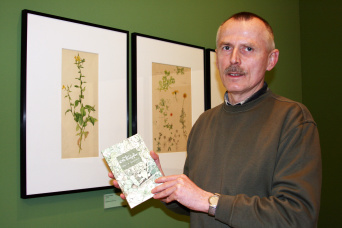 Ein Mann präsentiert ein Buch. Im Hintergrund eine Ausstellung mit Pflanzenabbildungen