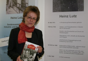 Dame mit rotem Schal steht vor einer Ausstellungstafel und hält ein Buch in den Händen.