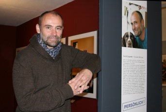 Ein Mann mit Vollbart und Halstuch lehnt an einer Ausstellungstafel, die sein eigenes Porträt abbildet.