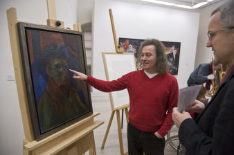 Ein lachender Mann zeigt auf ein Porträtbild. Rechts im Bild ein Zuschauer.