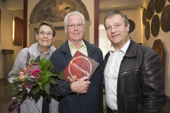 Eine Dame und zwei Herren, im Hintergrund sind Ausstellungsstücke des Museums zu sehen. Die Dame trägt einen Blumenstrauß, der Herr in der Mitte das geschenkte Buch.