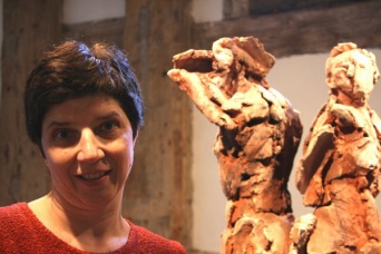 Eine Frau, im Bild links, blickt den Betrachter an. Rechts zwei Terrakottafiguren.