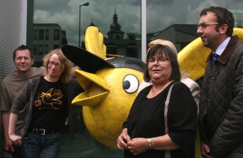 Ein gelber Vogel in Kugelform mit schwarzer Mütze. Links und rechts jeweils ein Mann und eine Frau.