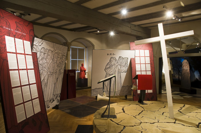 Installation im Geschichtslabor Vier große Tafeln, zwei mit Text auf rotem Grund und zwei mit Gelehrten schwarz auf weißem Grund. Rechts ein mannsgroßes Holzkreuz, schräg nach links kippend.