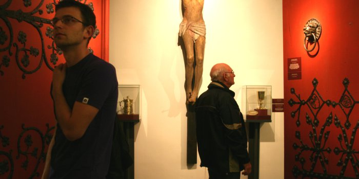 Im Zentrum des Bildes eine Holzplastik Jesus am Kreuz, links und rechts der Figur geöffnete rote Domtüren, rechts sieht man einen Türzieher aus dem Erfurter Dom, einen Löwenkopf.