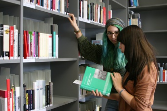 Zwei junge Frauen stehen, sich unterhaltend, in einer Bibliothek. Eine der beiden hält ein grünes Buch in der Hand.