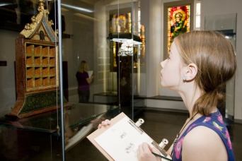 Ein Mädchen, rechts im Bild, schaut auf eine Vitrine, die sich links befindet. In der Hand hält sie ihre Zeichnung des abgebildeten Ausstellungsexponates.