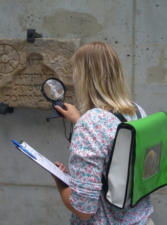 Mädchen mit grünem Rucksack und Lupe schaut auf einen jüdischen Grabstein.