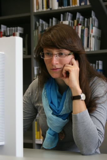 Eine junge Frau mit blauem Schal sitzt in einer Bibliothek und schaut auf einen Computerbildschirm. Im Hintergrund Bücherregale.