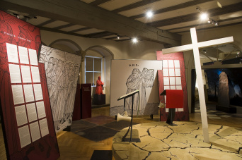Vier große Tafeln, zwei mit Text auf rotem Grund und zwei mit Gelehrten schwarz auf weißem Grund. Rechts ein mannsgroßes Holzkreuz, schräg nach links kippend.