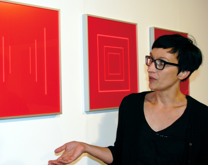 Eine Dame in schwarzer Kleidung, mit schwarzer Brille und rotem Lippenstift erklärt drei rote Kunstwerke, die sich im Hintergrund befinden.