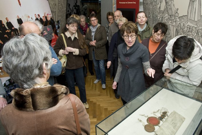 Zahlreiche Personen stehen um eine Vitrine. Die Kuratorin erläutert den Vitrineninhalt.