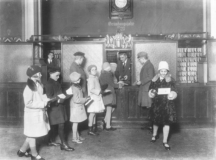 Sechs Kinder mit ihren Sparbriefen an einem Schalter. Über dem Schalter eine Bildillustration und ein Schriftzug "Gründungsjahr 1923"