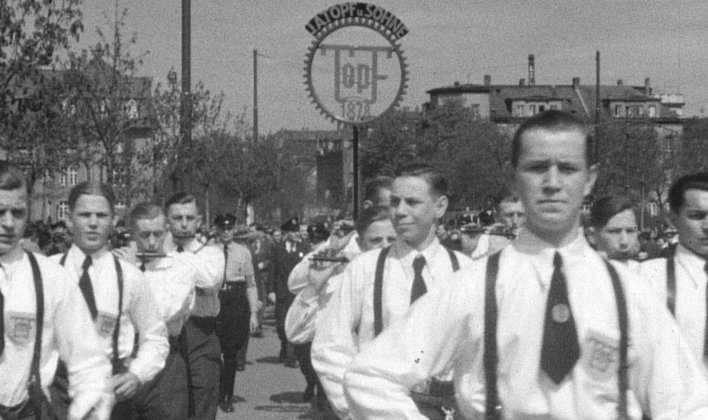 Junge Männer mit weißen Hemden und Krawatten marschieren in Formation. Im Hintergrund Uniformierte.