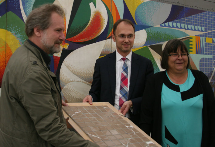 Drei Personen stehen um eine leicht erhöhte Mosaikplatte. Dahinter das Modell des Mosaikbildes.