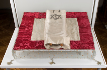 Foto: Torarolle im Toramantel (Inv. Nr. S 22b) auf Pultdecke (Inv. Nr. S 29) © Ulrich Fischer Auf einem Pult eine rot umrandete Decke, darauf die Torarolle, die in einer beigen Hülle liegt.