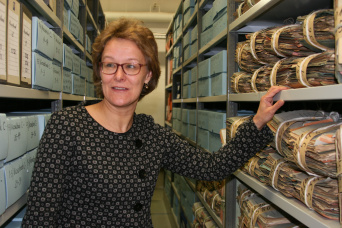 Eine Frau mit grauer Jacke und Brille steht zwischen zwei Regalreihen. In den Regalböden befinden sich Archiv-Schachteln mit der Aufschrift Vieselbach und gebündelte Akten.
