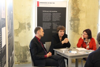 Drei Personen sitzen an einem Tisch. Im Hintergrund und links Ausstellungstafeln.