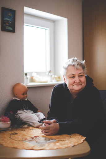 Ältere Dame mit grauem Haar sitzt an einem Tisch. Links auf dem Tisch eine Puppe. Im Hintergrund ein Fenster.