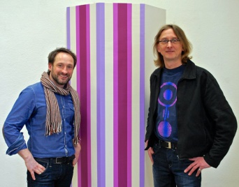 Zwei Herren, im Mittelpunkt eine farblich mit Streifen gestaltete Säule.