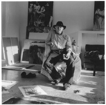 Ein Mann mit Hut sitzt auf einem Sessel, der mit einem Tuch belegt ist. Um ihn herum sind seine Arbeiten platziert. 