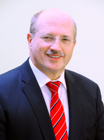 Ein Herr mit Brille und Schnurrbart, in dunkelblauem Anzug und roter Krawatte, im Viertelprofil.