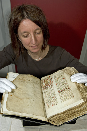 Eine Frau, mit weißen Handschuhen, hält eine alte Handschrift dem Betrachter entgegen.