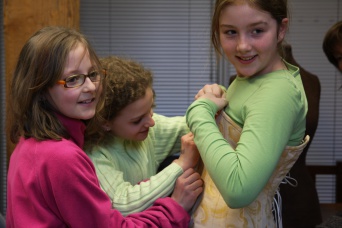 Ein Mädchen mit grünem Shirt bei der Anprobe eines historischen Kostüms. Zwei andere Mädchen helfen dabei.
