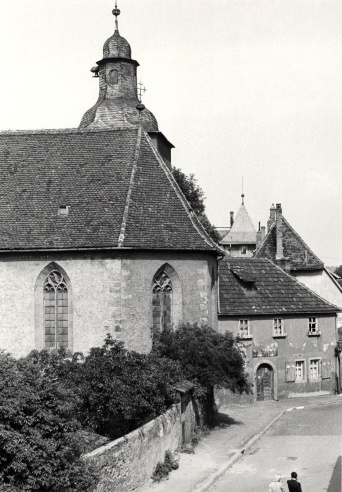 Links Kirchenschiff mit Kirchhof, rechts ein Straßenzug. Schwarz-Weiß-Aufnahme.