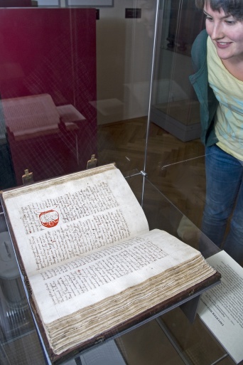 Frau, rechts im Bild, schaut auf eine Vitrine, in der eine alte Handschrift liegt.