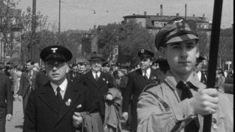 Mann mittleren Alters mit Brille und Mantel auf dem Arm bei einer Demonstration. Im Vordergrund rechts junger Mann als Fahnenträger.