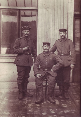 Drei Soldaten in Unform des württembergischen Infanterie-Regiments 124 auf einer braun gefärbten Fotografie. Der mittlere Soldat sitzt auf einem Stuhl, währenddessen die beiden anderen Soldaten rechts und links davon stehen. Im Hintergrund eine Hauswand mit Fenstern.