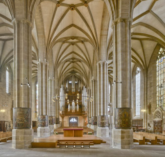 Altarinsel im Inneren einer Kathedrale, rechts und links davon mehrere Pfeiler, auf denen sich im jeweils unteren Teil halbrunde Gemälde befinden. 