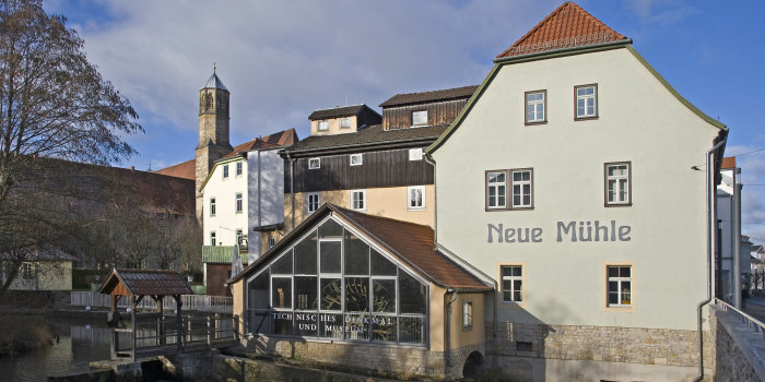 Mühlengebäude mit Glasfront, hinter der das große Mühlenrad zu sehen ist. Links ein Kirchturm.