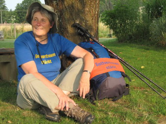 Pilgerin im Schneidersitz mit blauem T-Shirt mit der Aufschrift "Gottvertrauen stärkt" und Hut, rechts der Pilgerrucksack mit gleicher Aufschrift.