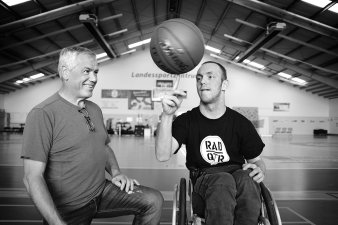 Schwarz-weißes Bild mit zwei Männer, einer davon im Rollstuhl,  in einer Sporthalle