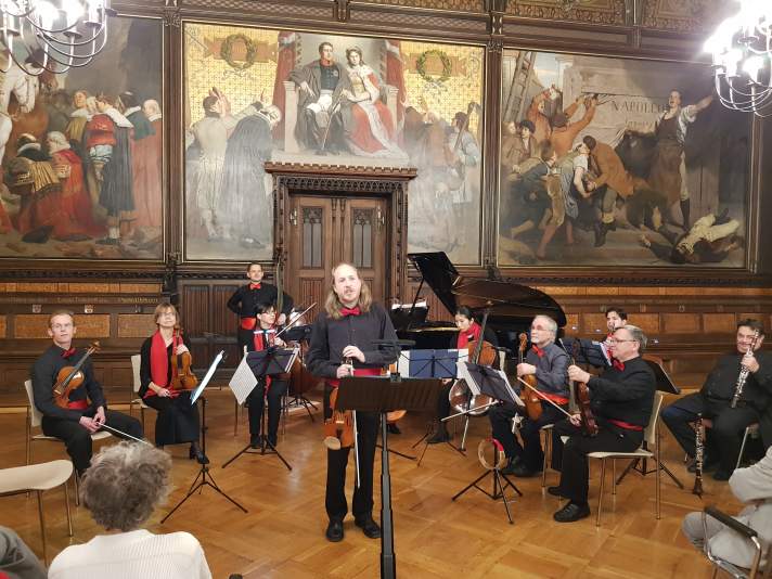 Ein Mann steht im Festsaal am Rednerpult, dahinter sind neun Musiker mit ihren Instrumenten zu sehen