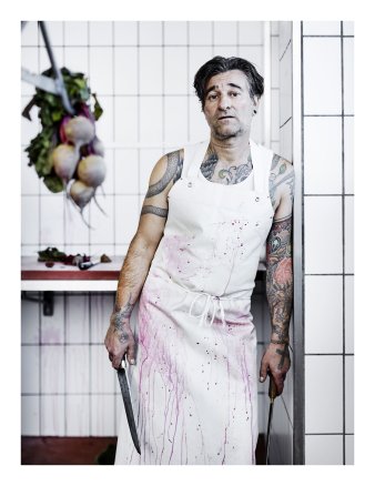 Ein Mann mit blutbespritzter weißer Schürze und einem Messer in der Hand lehnt am Türrahmen