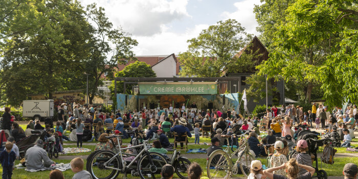 viele Menschen sitzen und stehen in einem Park vor einer Bühne