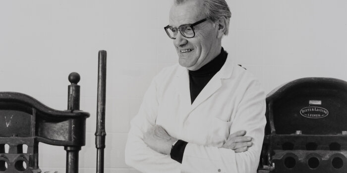 ein älterer Mann mit Brille und weißem Kittel steht vor Druckmaschinen