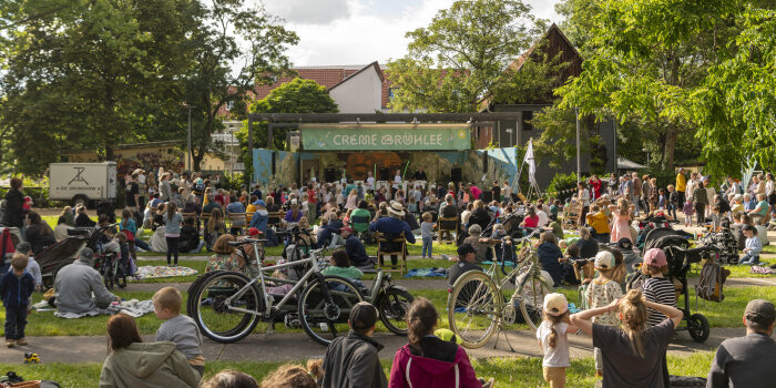 viele Menschen sitzen und stehen in einem Park vor einer Bühne
