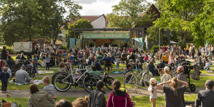 viele Menschen sitzen und stehen in einem Park vor einer Bühne
