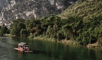 Im Hintergrund sind Berge, im Vordergrund ein Fluss mit einem Hausboot zu sehen. Das Bild zeigt eine chinesische Landschaft.