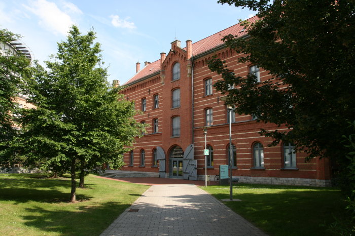 Benaryspeicher im Sparkassen-Finanzzentrum Ein Weg führt zum Tor eines großen, mit Querstreifen versehenen Hauses. Rechts und links Bäume, die die Wiese vorm Haus verschatten.