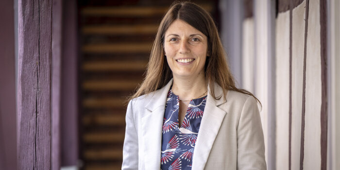 eine Frau mit langen braunen Haaren, gemusterter Bluse und hellgrauem Blazer