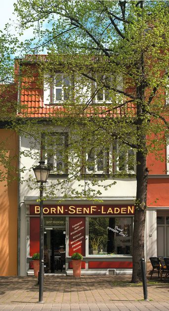 Foto: Born-Senf-Laden in Erfurt © Stadtverwaltung Erfurt Ein schmales Haus mit zwei Buchsbaum-Töpfen rechts und links vorm Eingang. Rechts vom Haus ein großer Baum. Über der Eingangstür und dem Schaufenster ein Schriftzug, auf dem Born-Senf-Laden steht. Links vor dem Haus eine historische Straßenlaterne