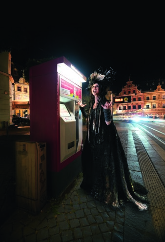 eine mit schwarzem Kleid und Kopfschmuck verkleidete Frau an einem Fahrkartenautomaten der Straßenbahn
