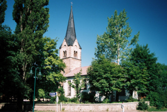 Kirche mit spitzem Turm inmitten grüner Bäume.