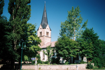 Evangelische St. Nikolauskirche, „Kirche am Pilgerweg“, im Evangelischen Kirchspiel Hochheim-Schmira Kirche mit spitzem Turm inmitten grüner Bäume.