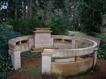 Ein in Kreisform gestaltete Grabanlage. Die kniehohe Balustrade aus Naturstein begrenz die Anlage und gestattet an einer Stelle den Zugang zum Innenraum, hier fügt sich eine Bank am Rand ein und das Grabmal ist als Quader eingefügt.